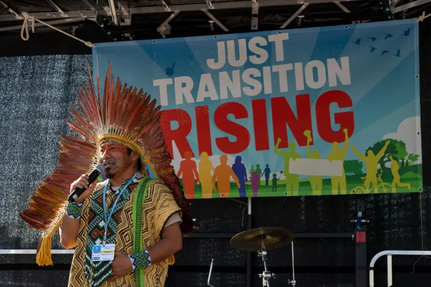 A man standing on a stage holding a microphone. Behind him is a colourful sign that reads: Just Transition Rising.