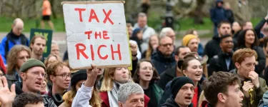Man holds a banner saying 'tax the rich'. He is surrounded by people.