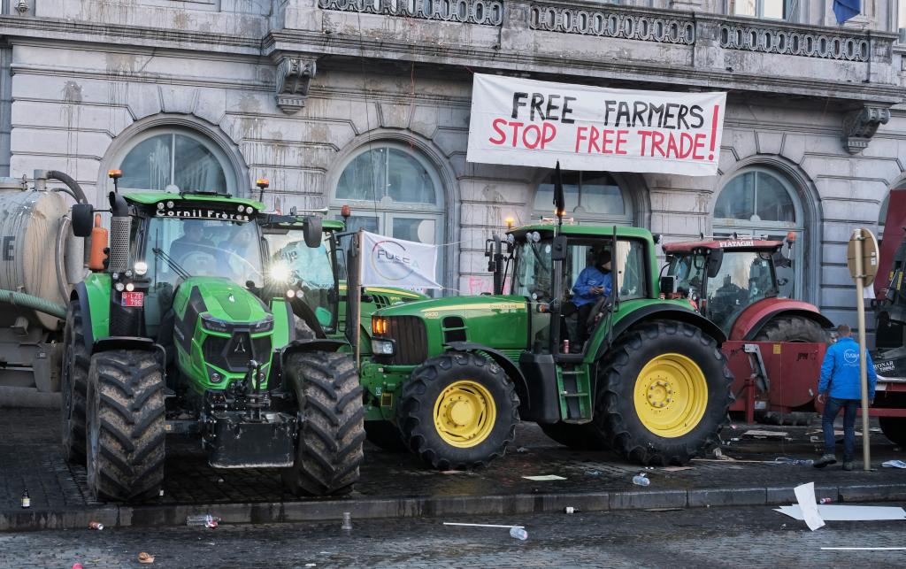 Green tractors parked in protest outside a building. Above the tractors is a banner which reads: "Free farmers, stop free trade!"