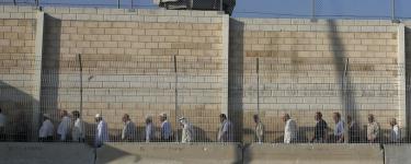 Palestinian workers in Israel protest dehumanising conditions at checkpoints. A queue of people stand in a line in front of a tall wall at a checkpoint. Source: Wikipedia