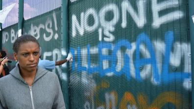 A black person standing in front of the fence at at a protest at Yarl's Wood Immigration Removal Centre. Graffiti on the fence reads: "No Borders! No one is illegal". Photo: IDJ Photography