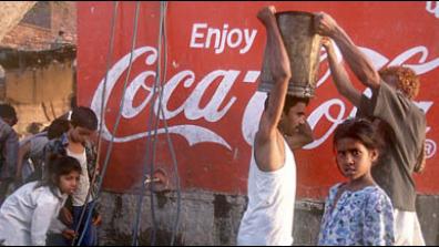People standing in front of a Coca-Cola advert. One person with a white vest an moustache is passing a large drum that he is carrying on his head to another person.